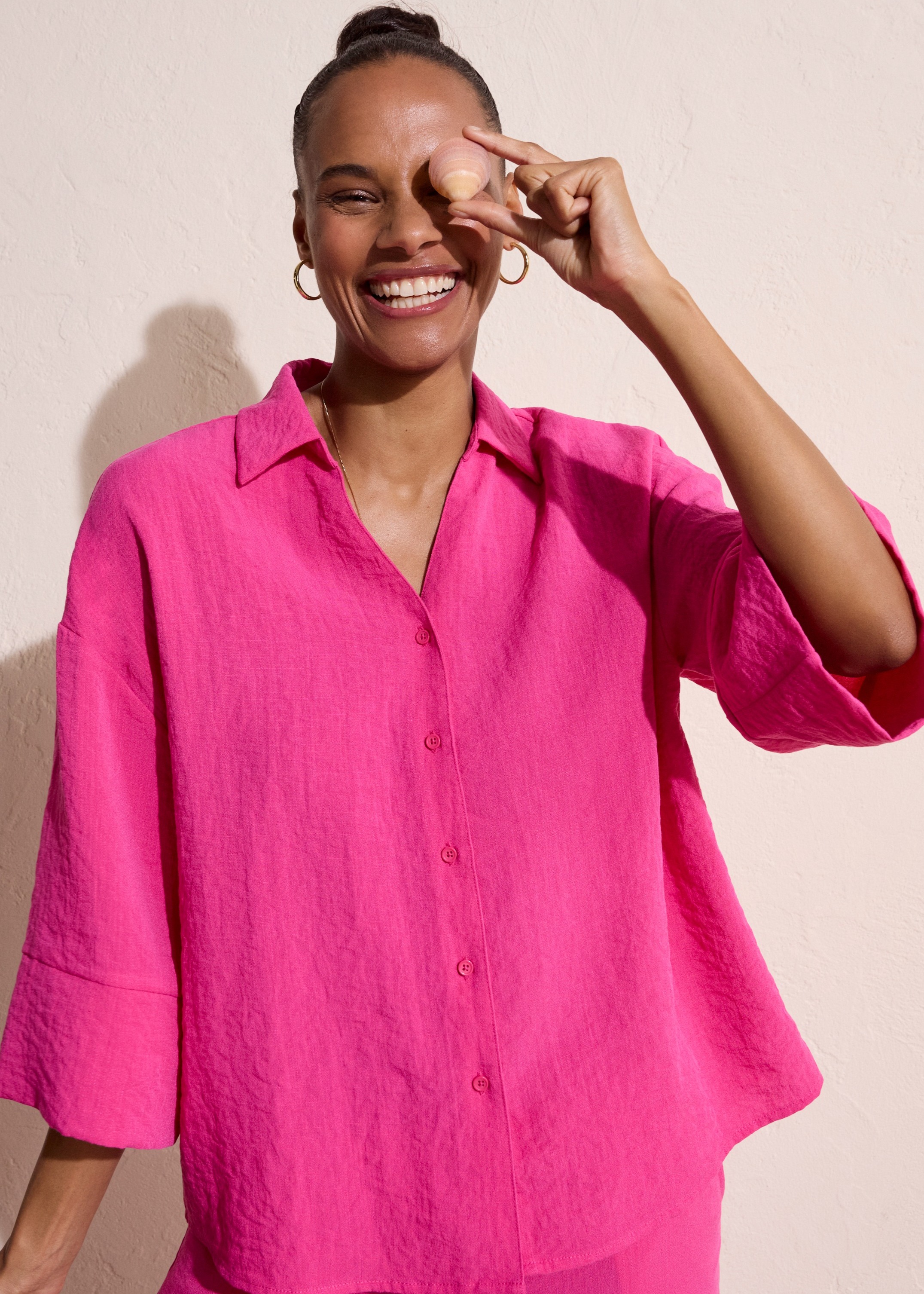 Eine lächelnde Frau trägt eine leuchtend pinke Bluse mit Dreiviertel-Ärmeln und klassischem Kragen.