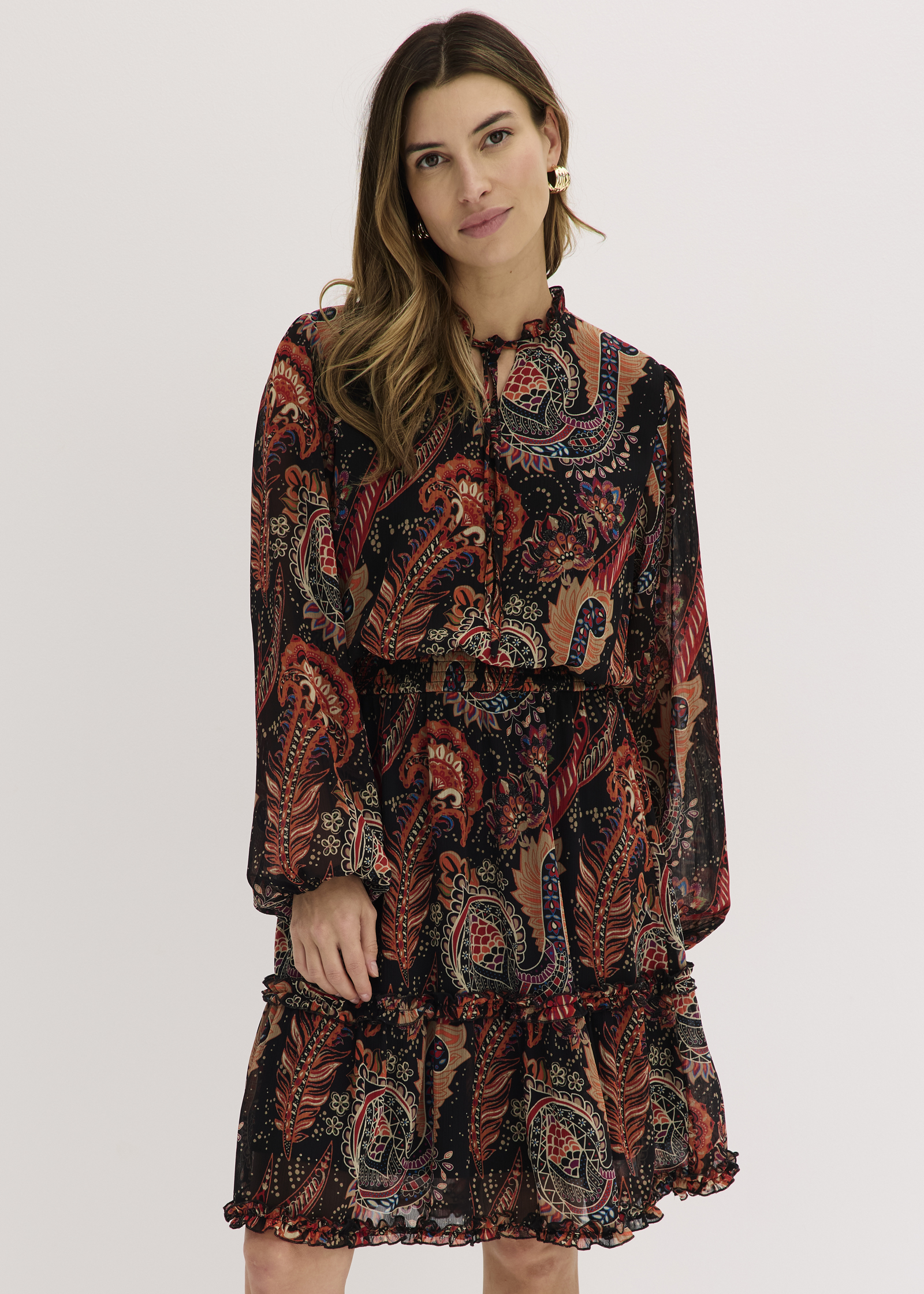 Sie trägt ein fließendes Kleid mit buntem Paisley-Muster und Rüschendetails.