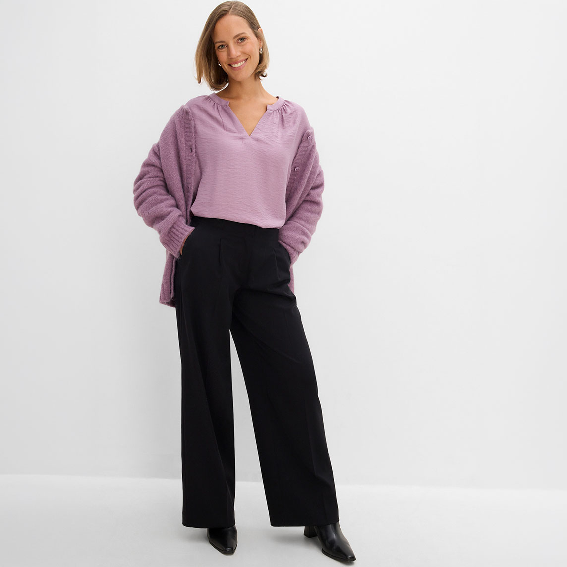 Sie trägt eine lila Bluse, dazu einen Strickcardigan und eine schwarze weite Hose.