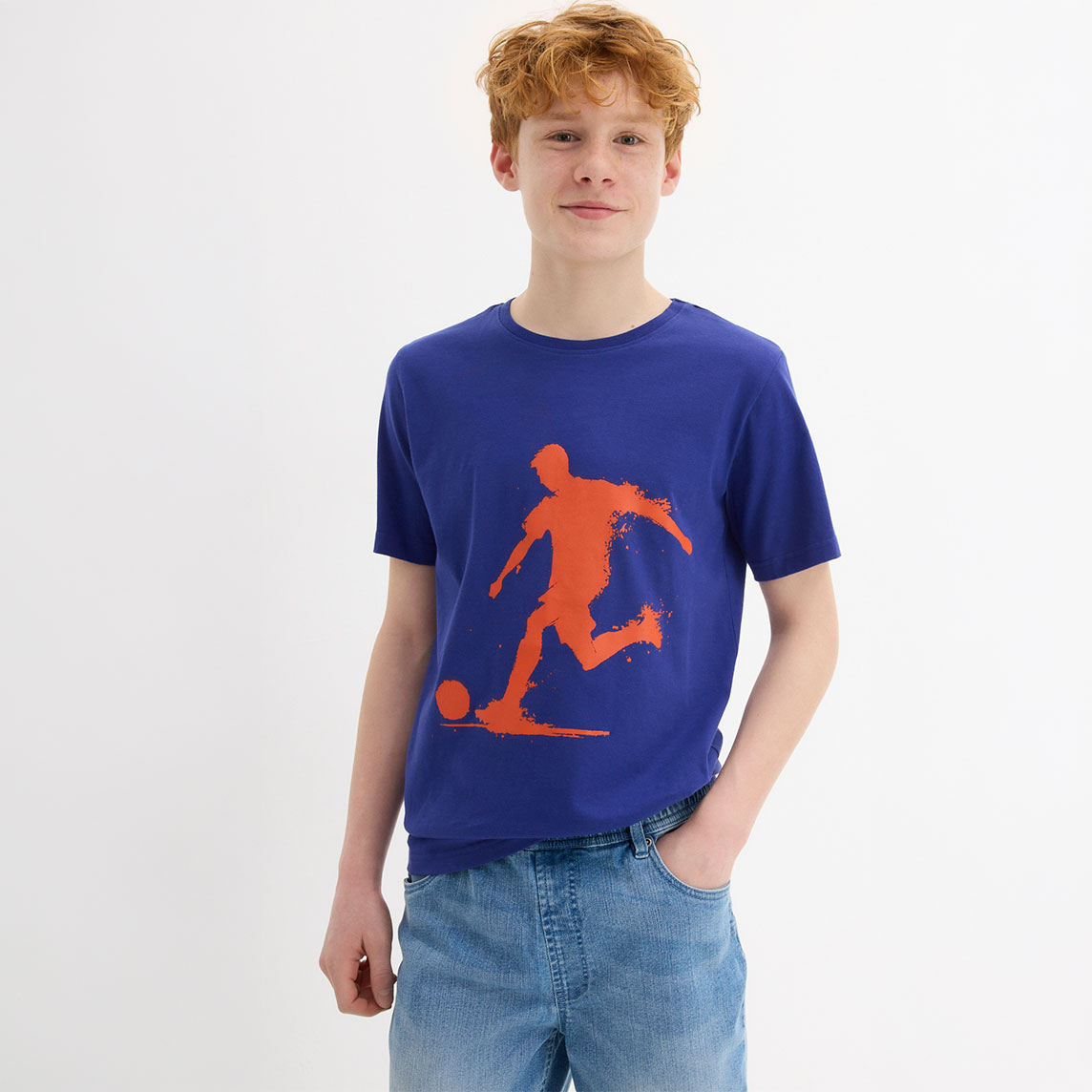 Junge trägt blaues T-Shirt mit orangem Fußball-Motiv und heller Jeans.