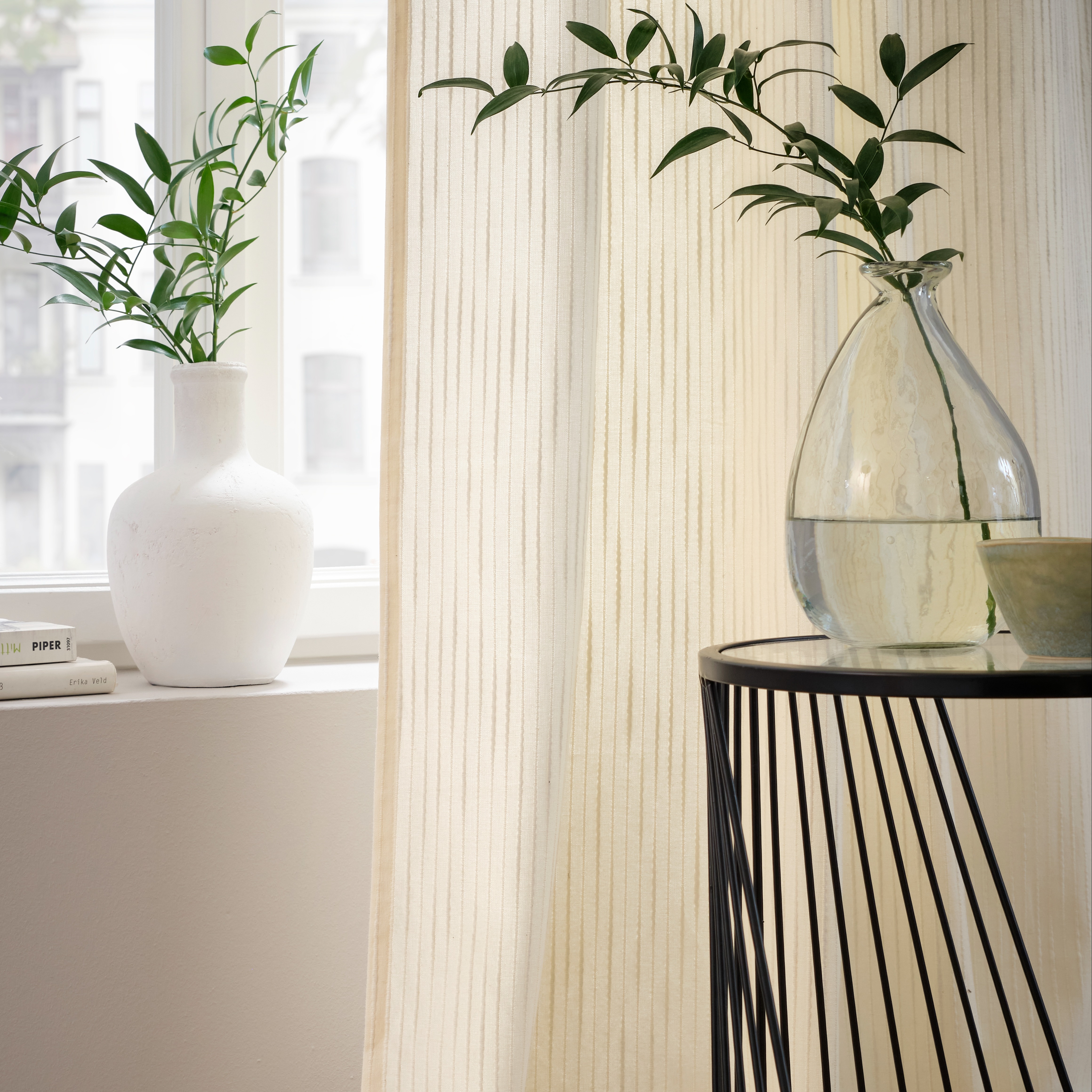 Ein Fenster mit cremefarbener Gardine, sowie Dekoration auf der Fensterbank und einem kleinen Tisch davor.