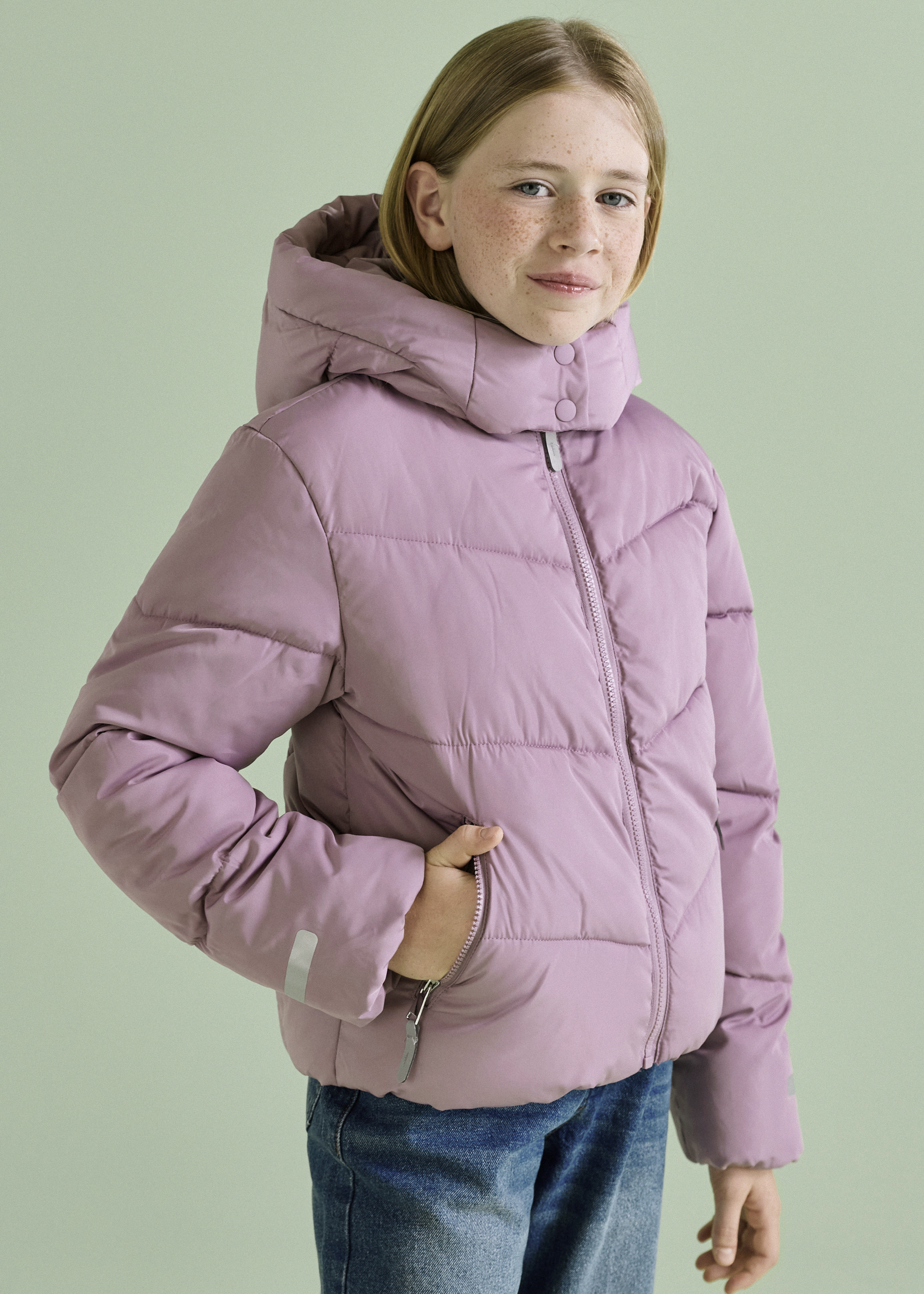 Dieses Mädchen trägt eine warme lila Steppjacke mit Kapuze und blaue Jeans.