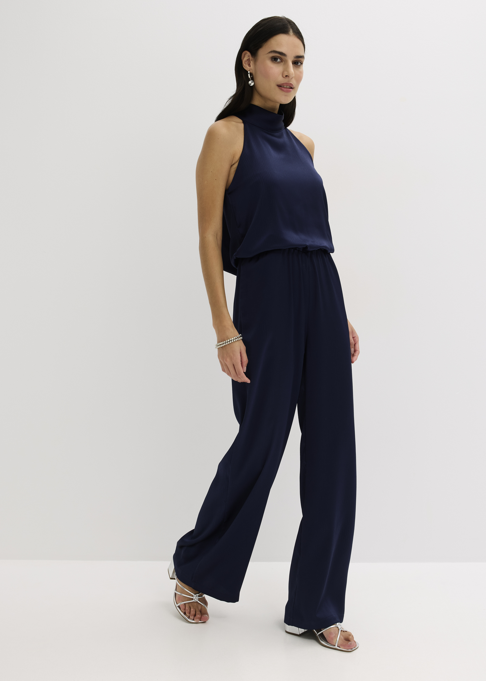 Eleganter, marineblauer Jumpsuit mit weitem Bein und hohem Kragen, kombiniert mit weißen Sandalen.