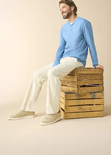 Er trägt ein hellblaues Langarmshirt, eine helle Hose und beige Sneaker.