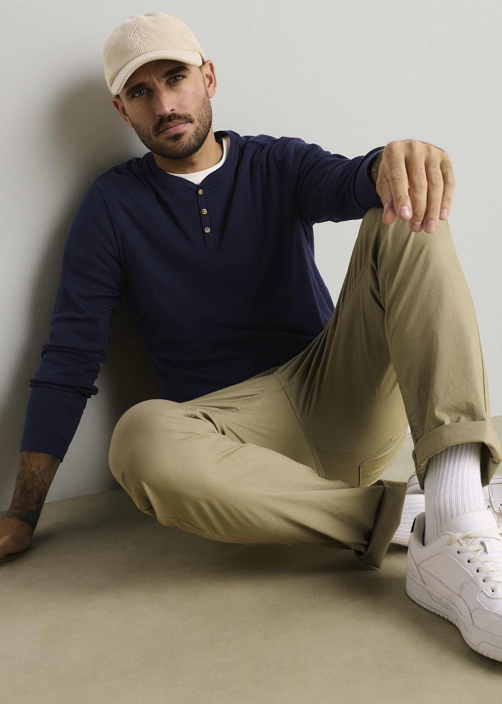 Beige Mütze, blaues Henley-Shirt, beigefarbene Hose und weiße Sneaker sind zu sehen.