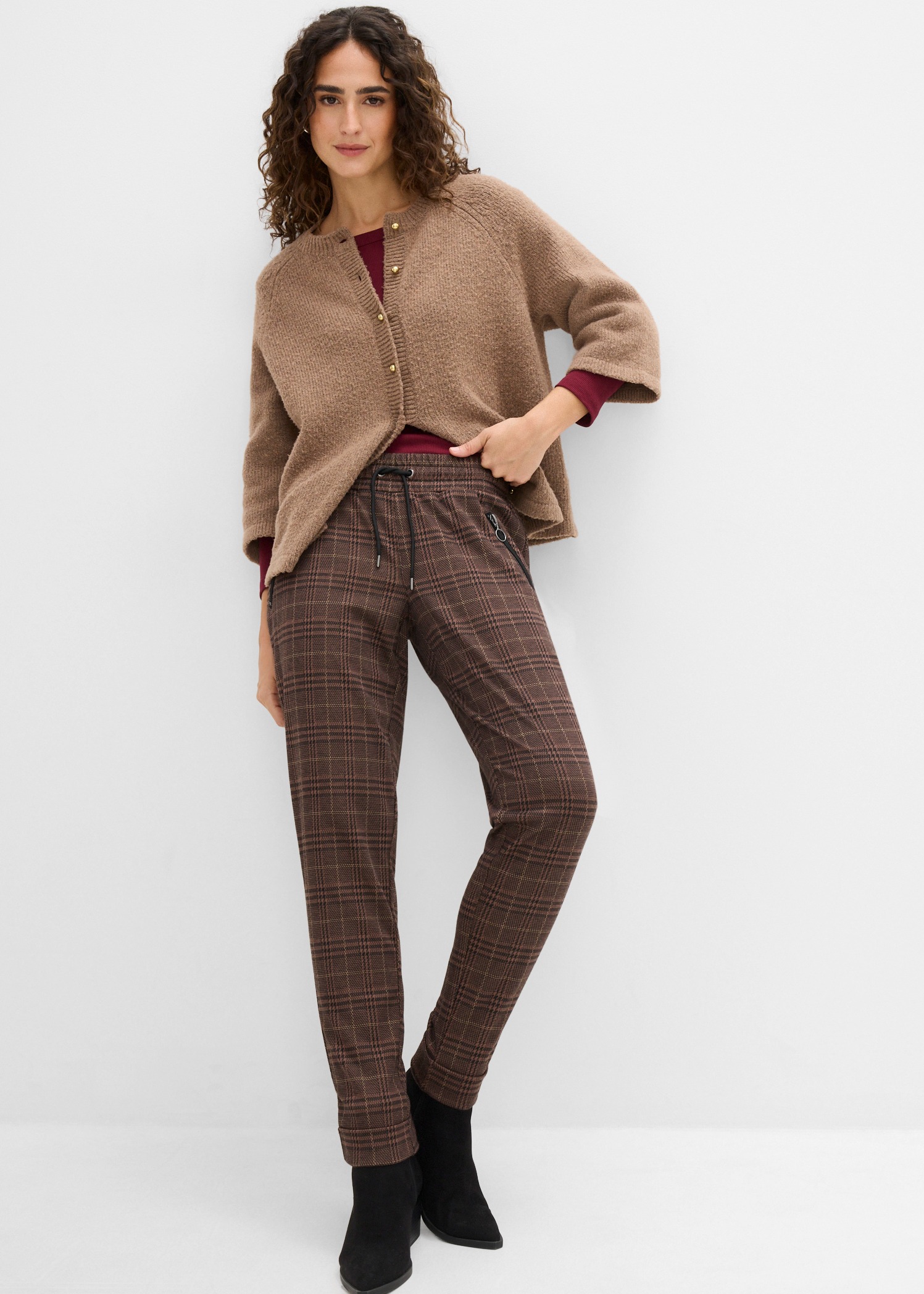 Braune Strickjacke, rotes Oberteil, karierte Hose und schwarze Stiefeletten.