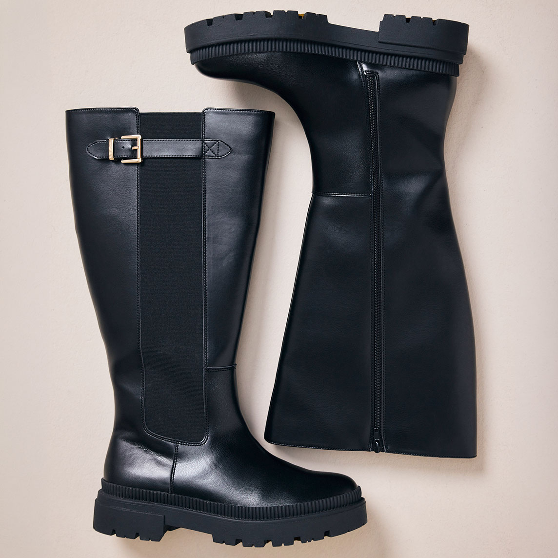 Zwei schwarze, kniehohe Stiefel mit markanter Profilsohle und Schnallendetail.