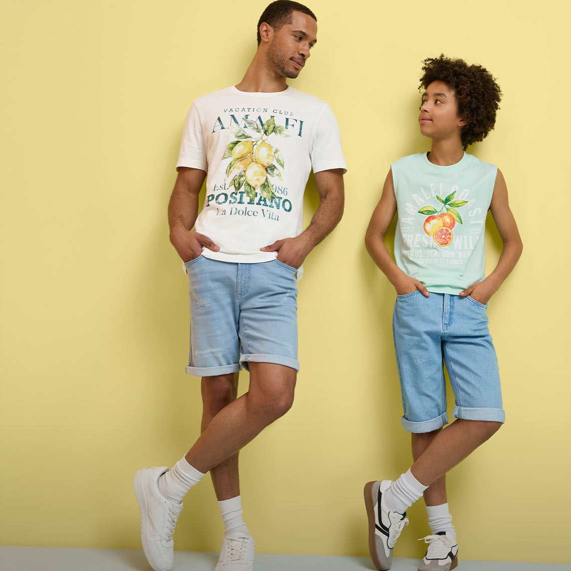 Zwei Personen tragen sommerliche Outfits mit bedruckten T-Shirts, Jeans-Shorts und weißen Sneakers.