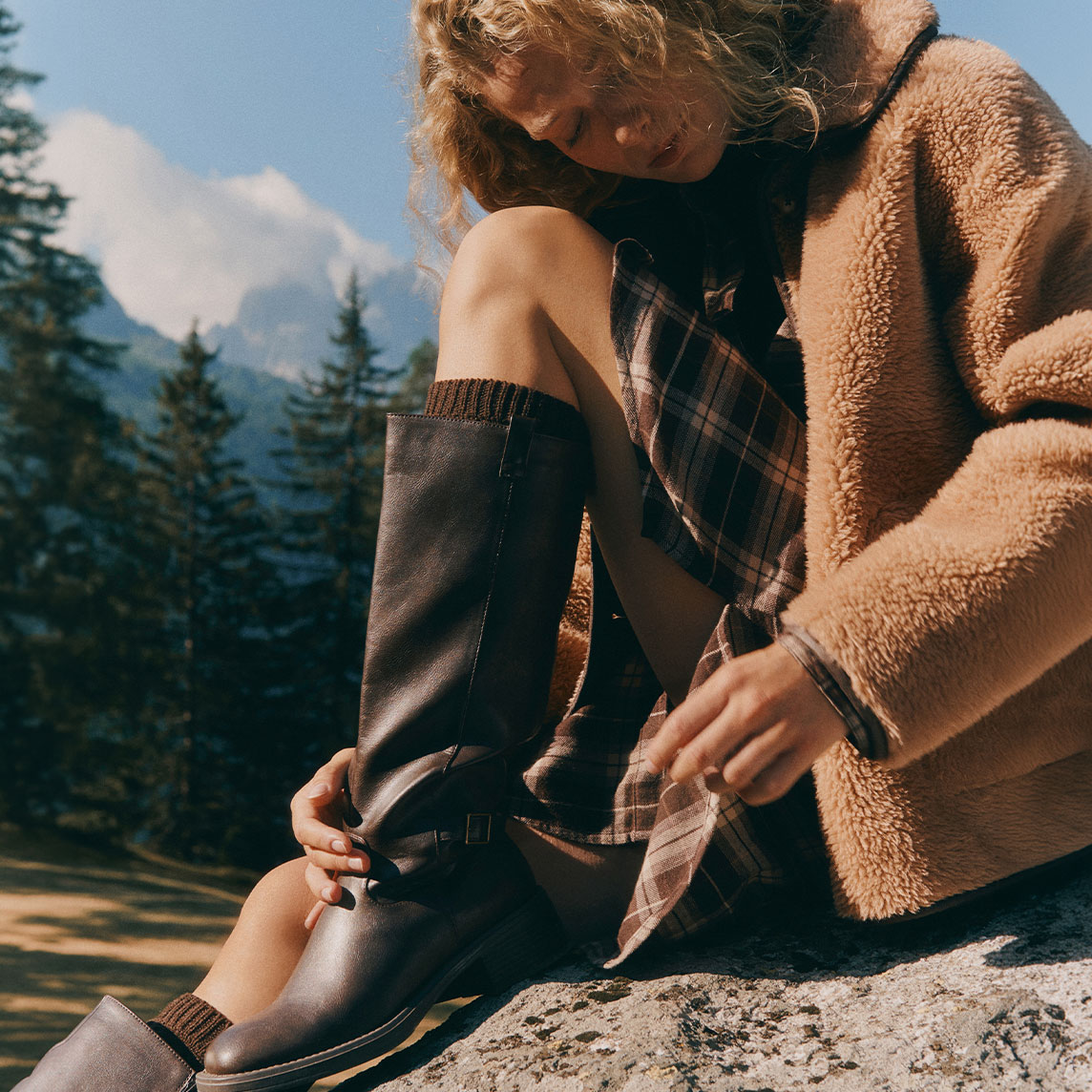 Kuschelige Teddyjacke, kariertes Kleid und braune Lederstiefel für Outdoor-Abenteuer.