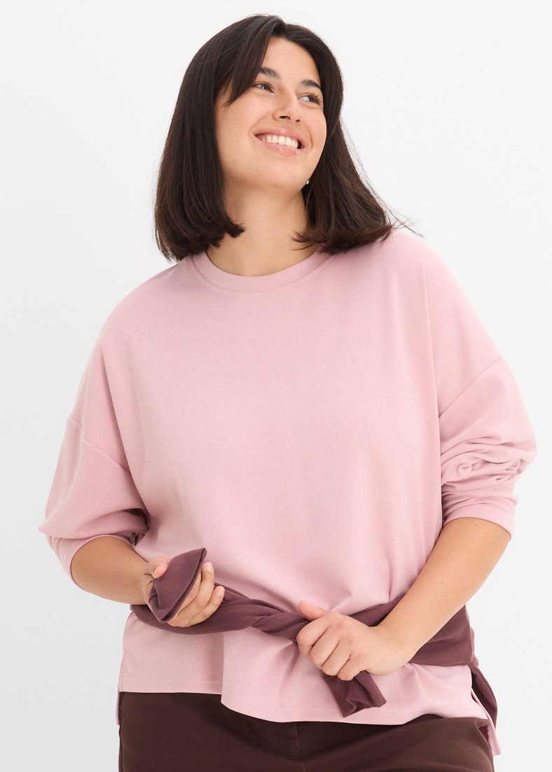 Rosa Oversize-Shirt, braune Hose und ein um die Taille gebundener Pullover.