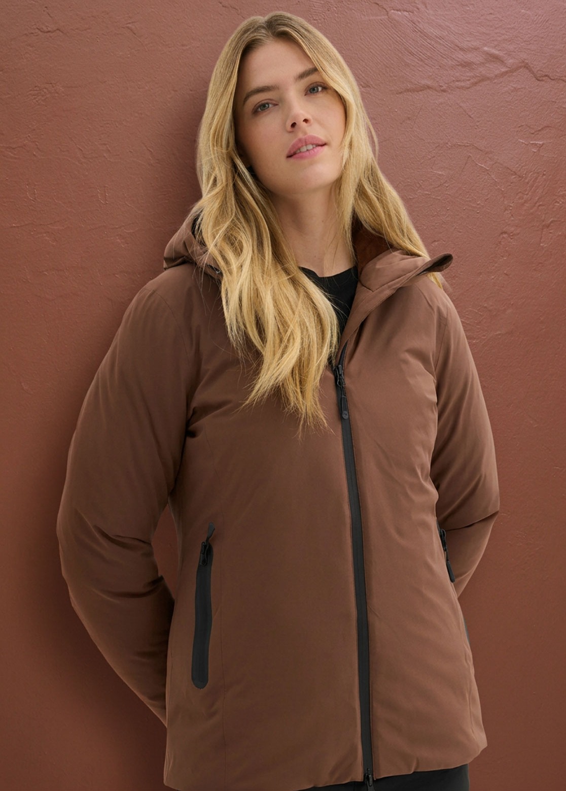 Sie trägt eine stilvolle, braune Steppjacke mit Kapuze und schwarzen Reißverschlüssen.