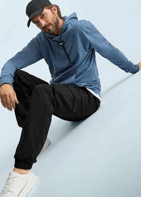 Der Mann trägt einen blauen Kapuzenpullover, schwarze Cargohose, weiße Sneaker und Kappe.