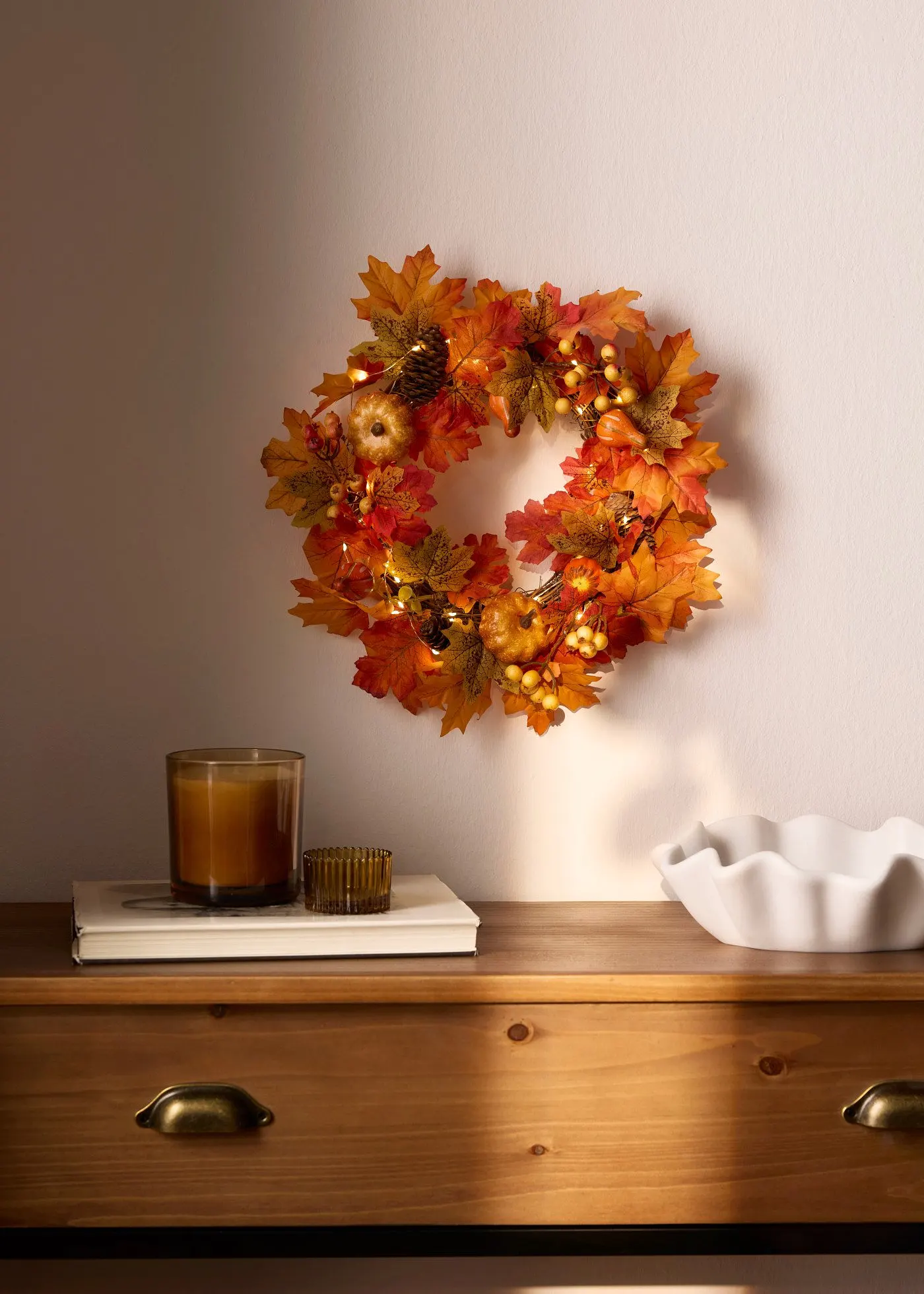 Wand ziert ein beleuchteter Herbstkranz. Kerzenlicht und eine Schale auf dem Holztisch.
