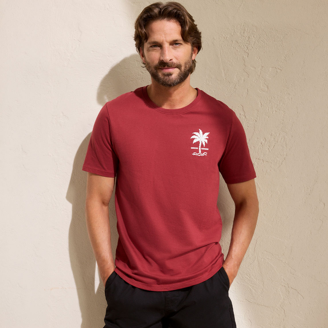Ein Mann trägt ein rotes T-Shirt mit kleinem weißen Logo und schwarze Hose.