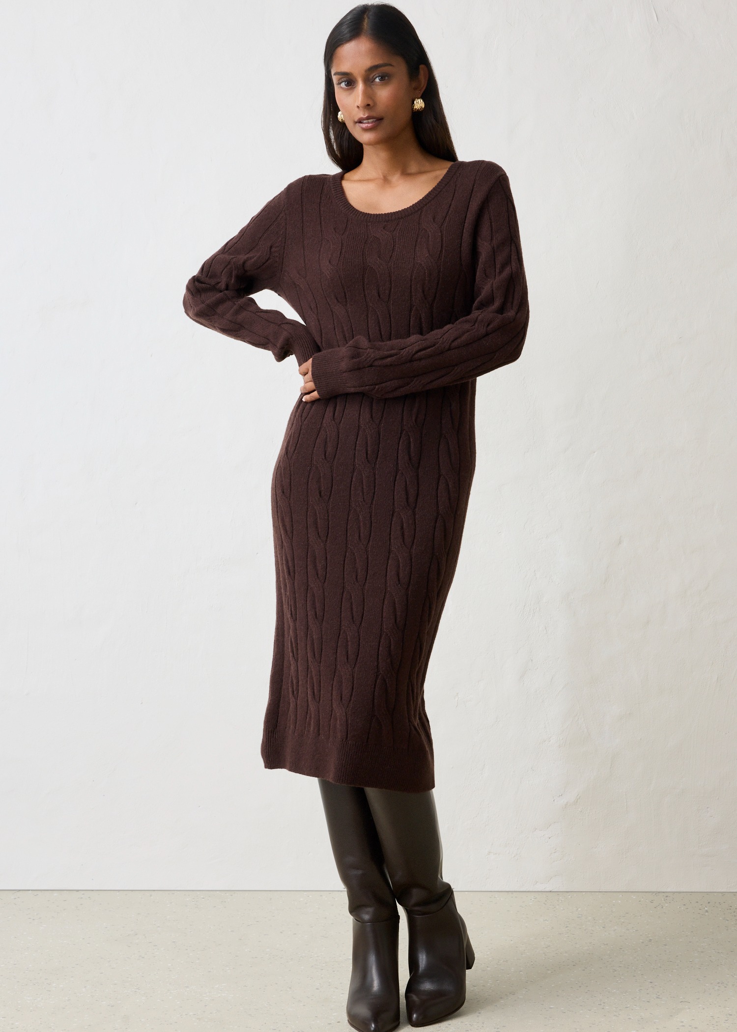 Elegantes braunes Strickkleid mit Zopfmuster, kombiniert mit hohen dunklen Stiefeln und Ohrringen.