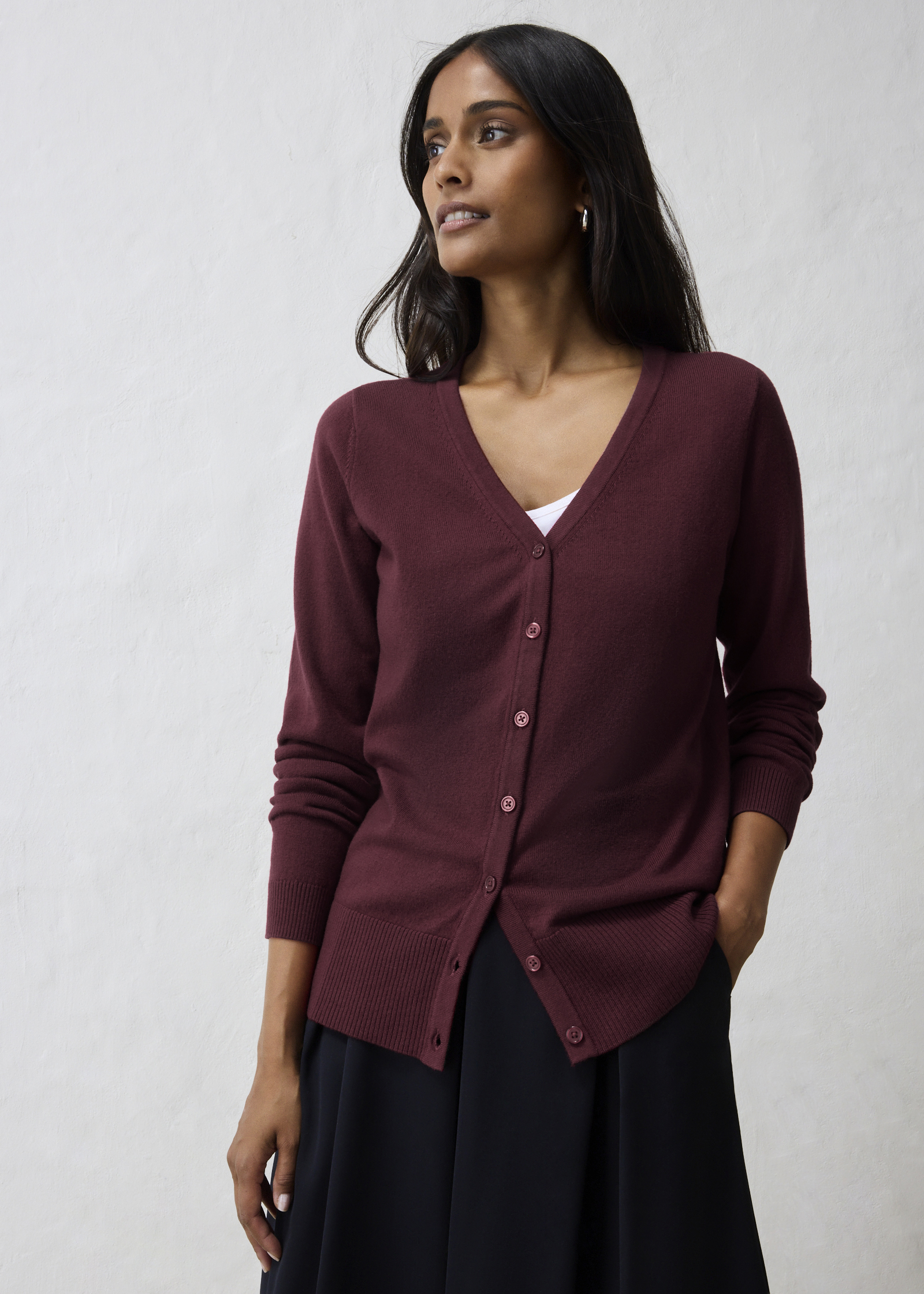 Die Frau trägt eine stilvolle, bordeauxfarbene Strickjacke, ein weißes Top und einen dunklen Rock.