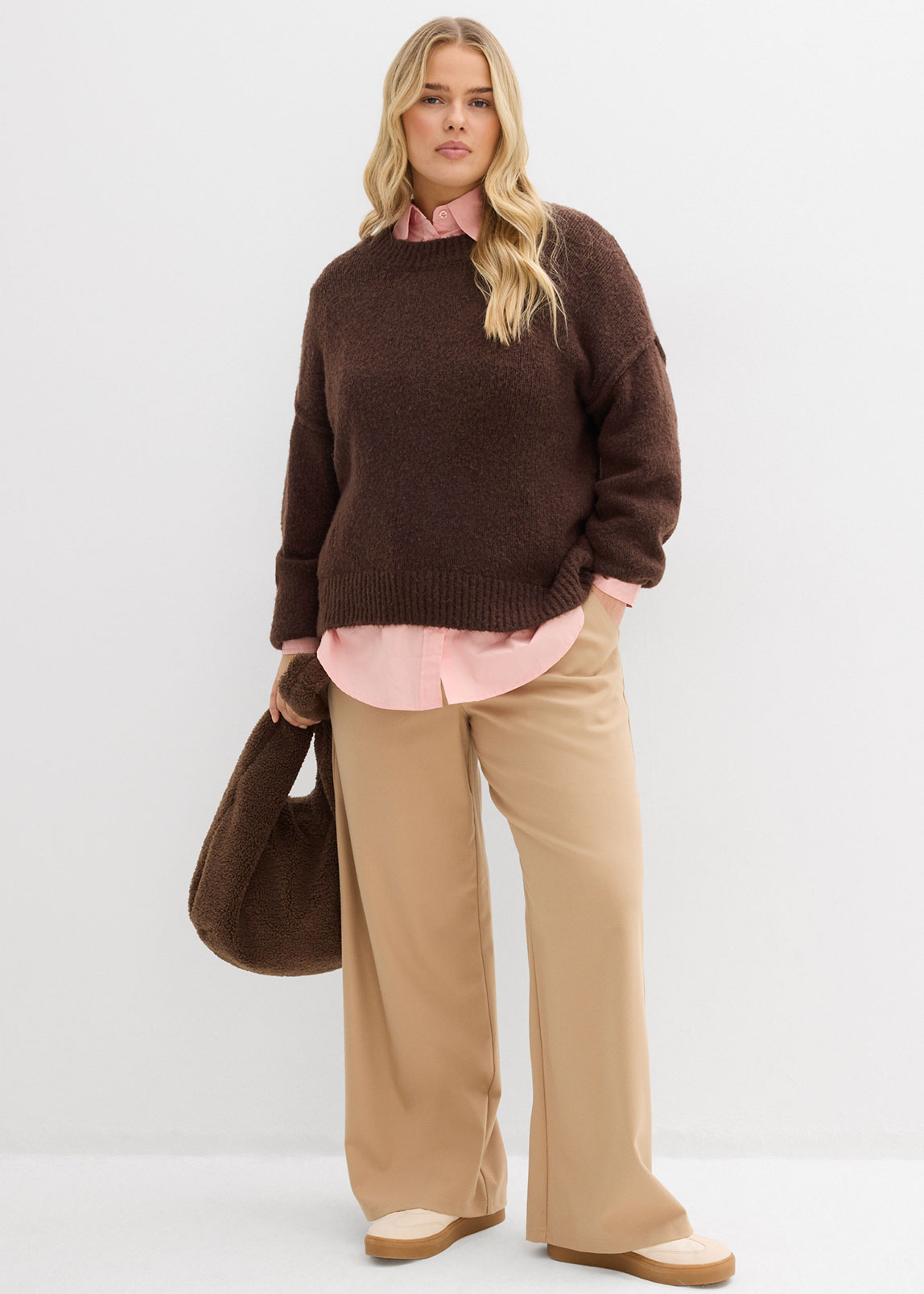 Brauner Pullover über einem rosa Hemd, beige Hose und braune Handtasche.