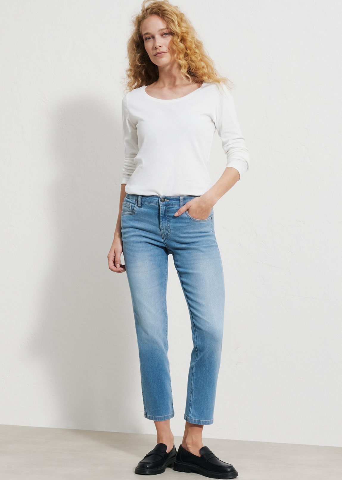 Sie trägt ein weißes Langarmshirt, eine hellblaue Jeans und schwarze Loafer.