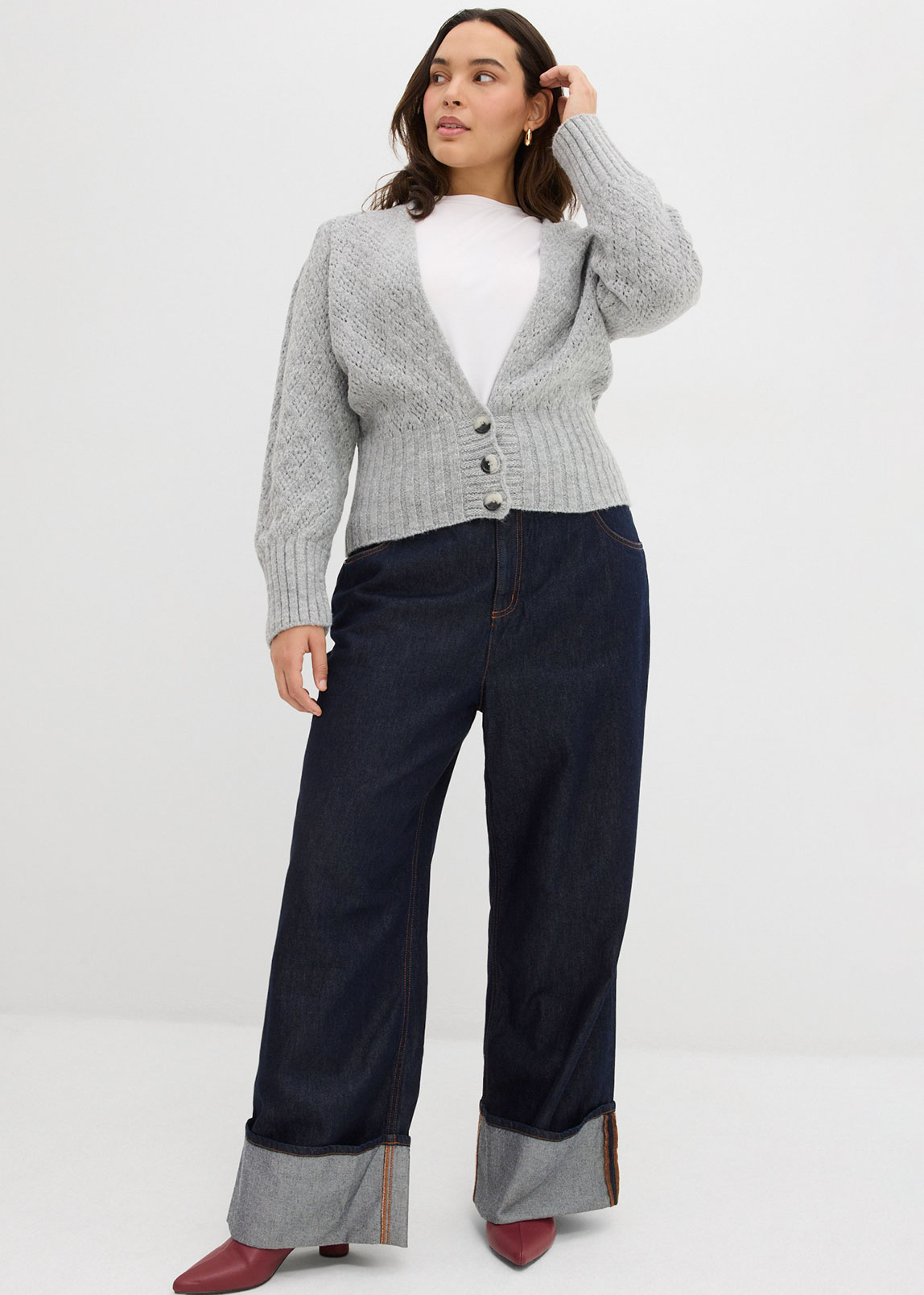 Grauer Strickcardigan über weißem Top, dunkle Jeans mit Umschlag und rote Schuhe.