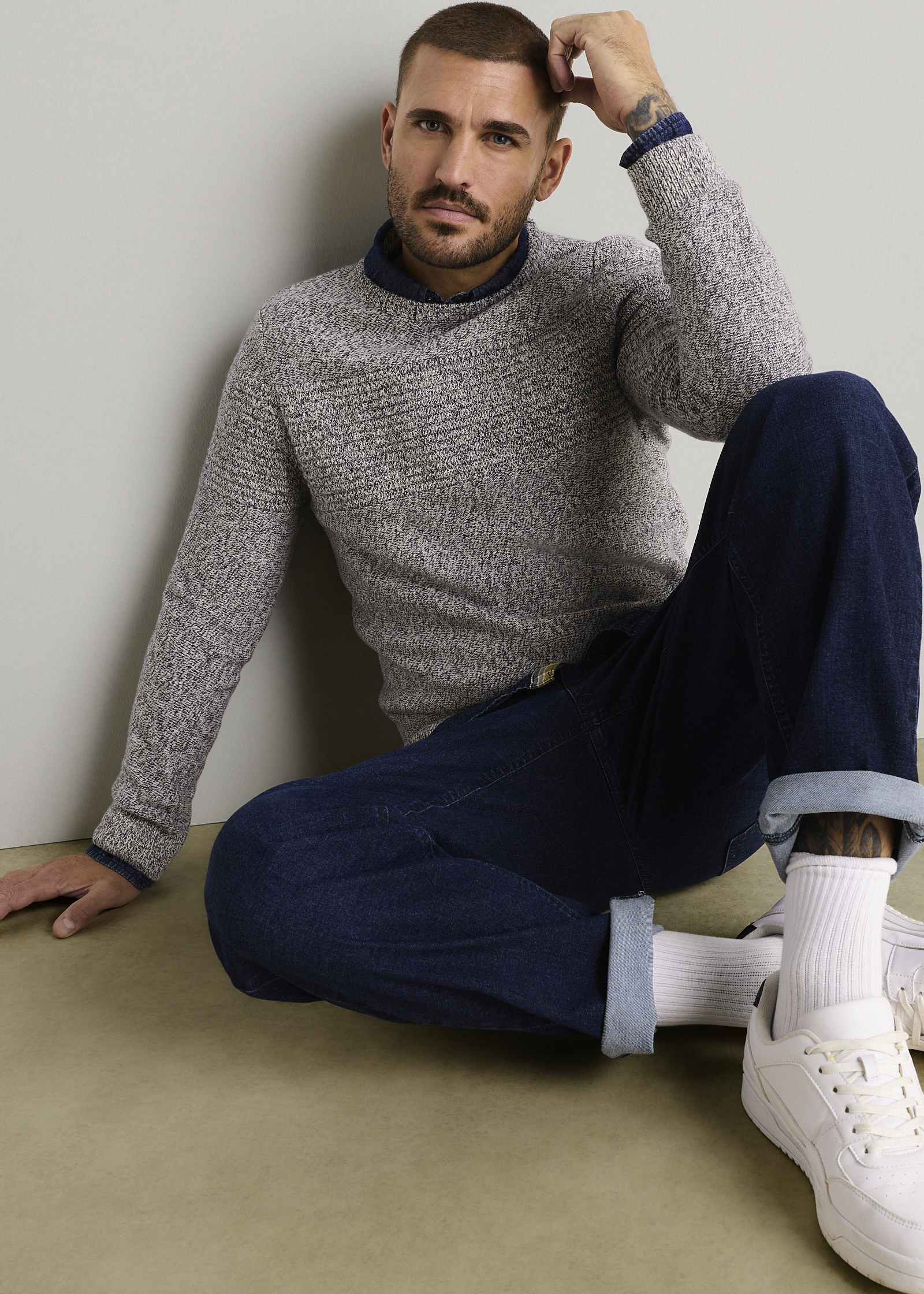 Grauer Strickpullover über blauem Hemd, dunkle Jeans, weiße Socken und Sneaker.
