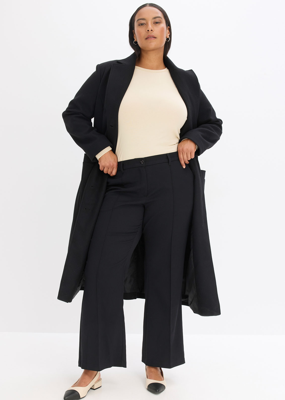 Schwarzer Mantel über cremefarbenem Pullover, kombiniert mit weiter schwarzer Hose.