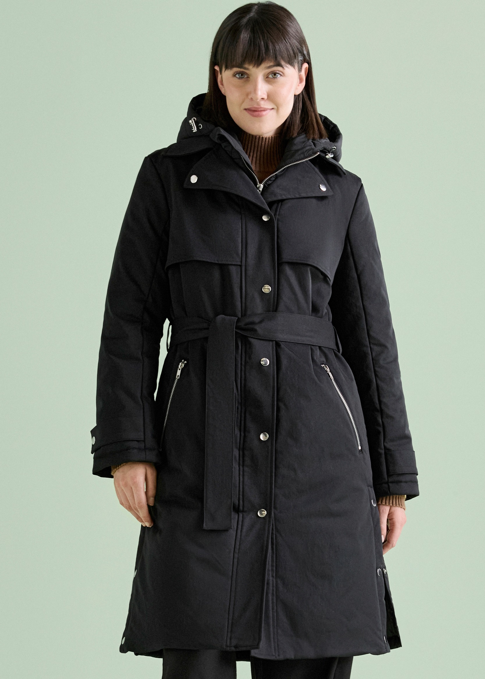 Schwarzer, eleganter Wintermantel mit Gürtel, Kapuze und Reißverschlussdetails an einer Frau.
