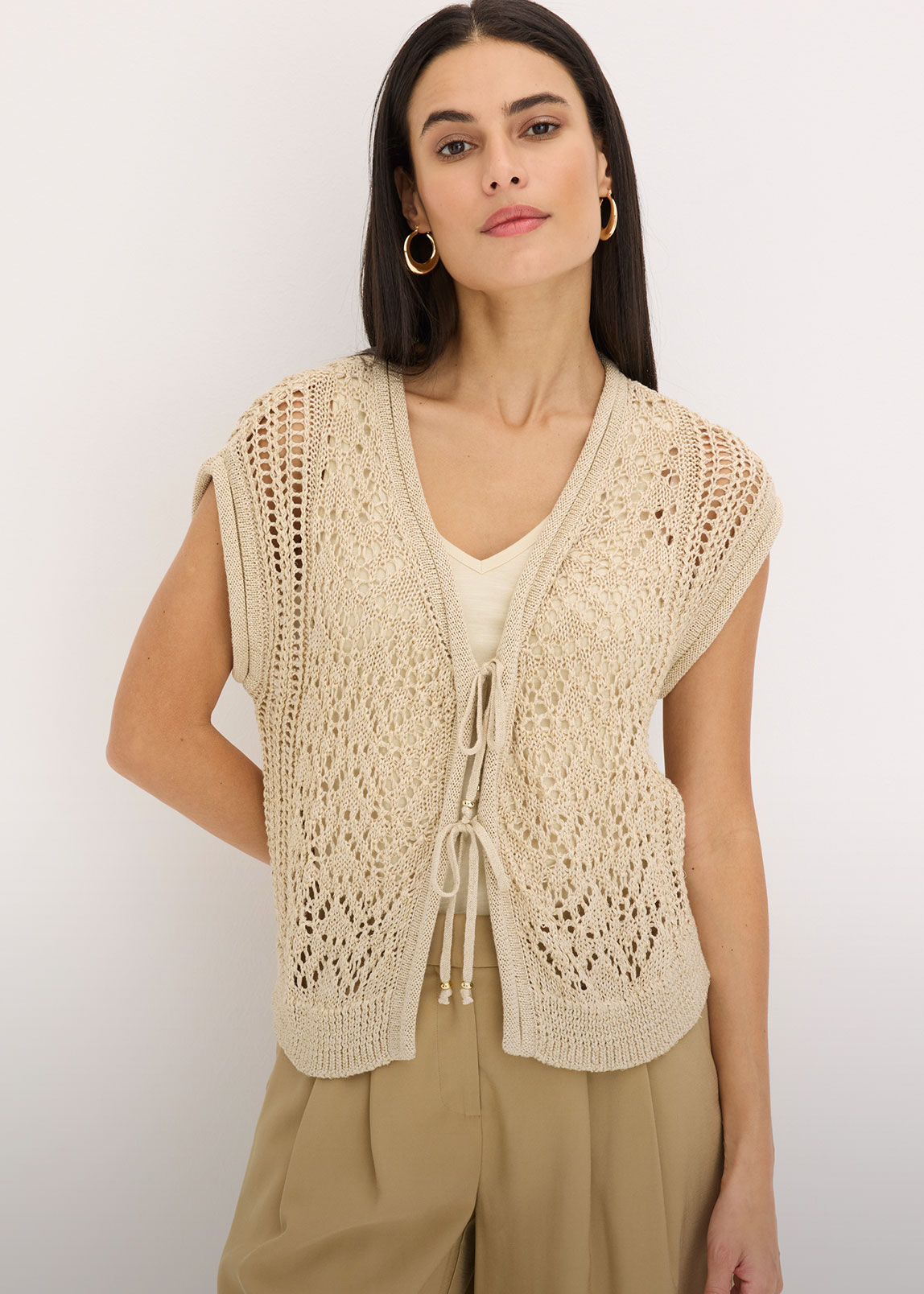Beigefarbene, ärmellose Ajour-Strickweste mit Bindedetails über hellem Top und weiter Hose.