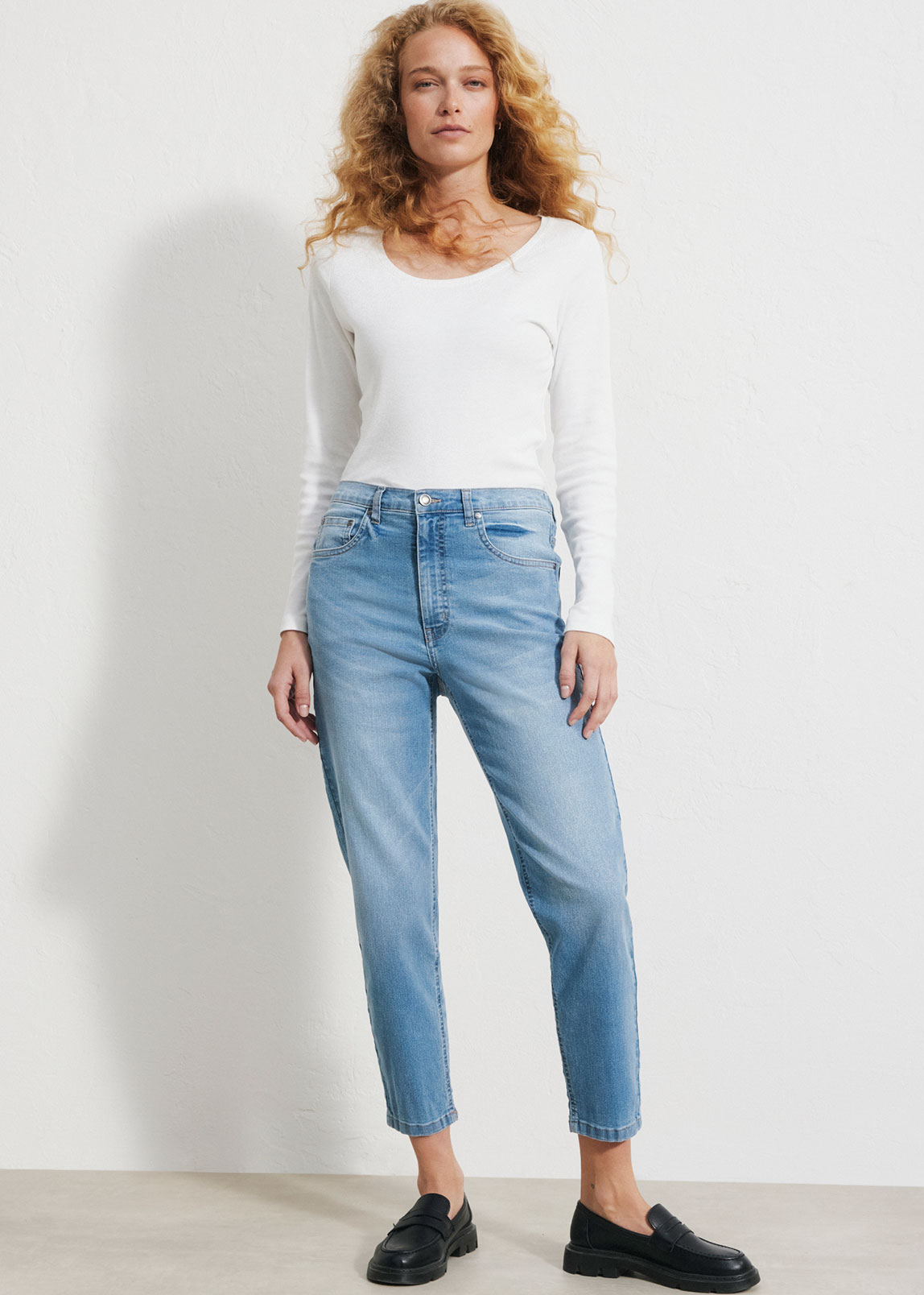 Die Frau trägt ein weißes Langarmshirt, hellblaue Jeans und schwarze Loafer.