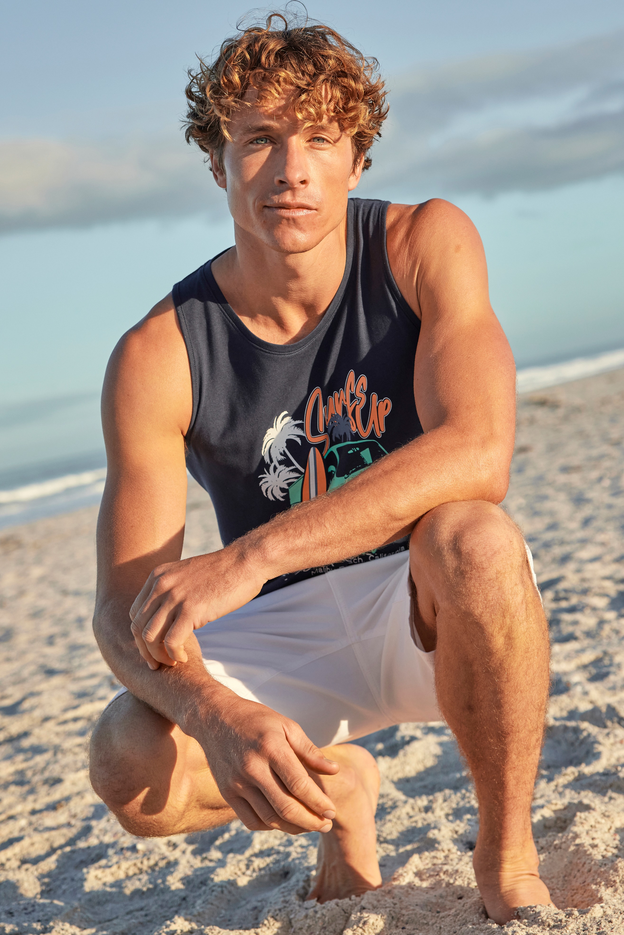 Mann trägt ein blaues Tanktop mit Motiv und weiße Shorts am Strand.