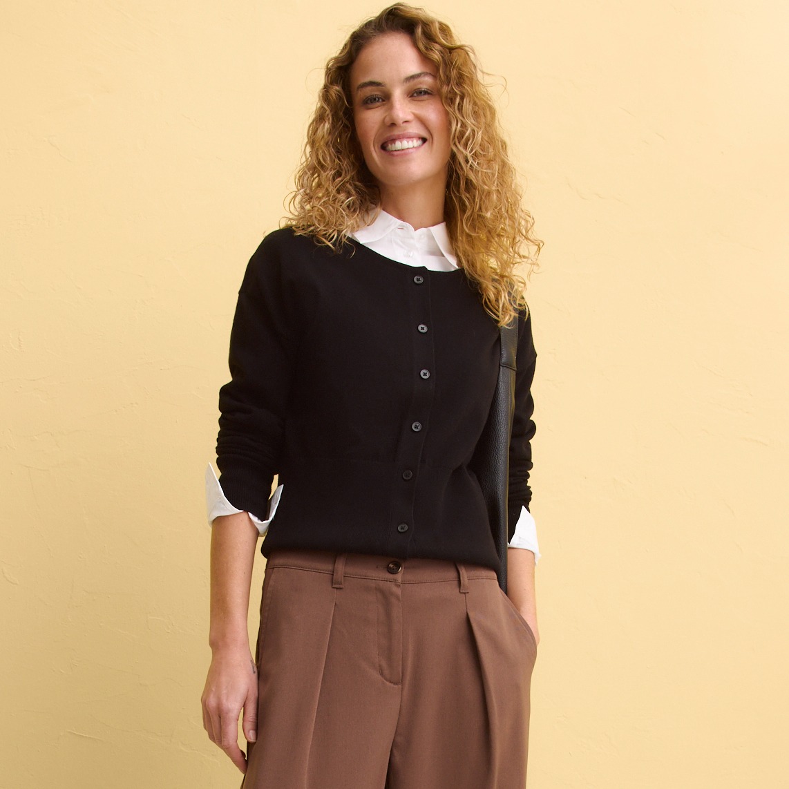 Schwarze Strickjacke mit Knöpfen über weißer Bluse, kombiniert mit brauner Hose.