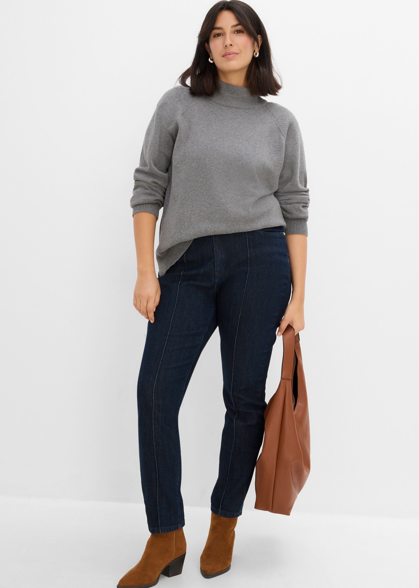 Grauer Stehkragenpullover, dunkle Jeans mit Nähten, braune Stiefeletten und Tasche.
