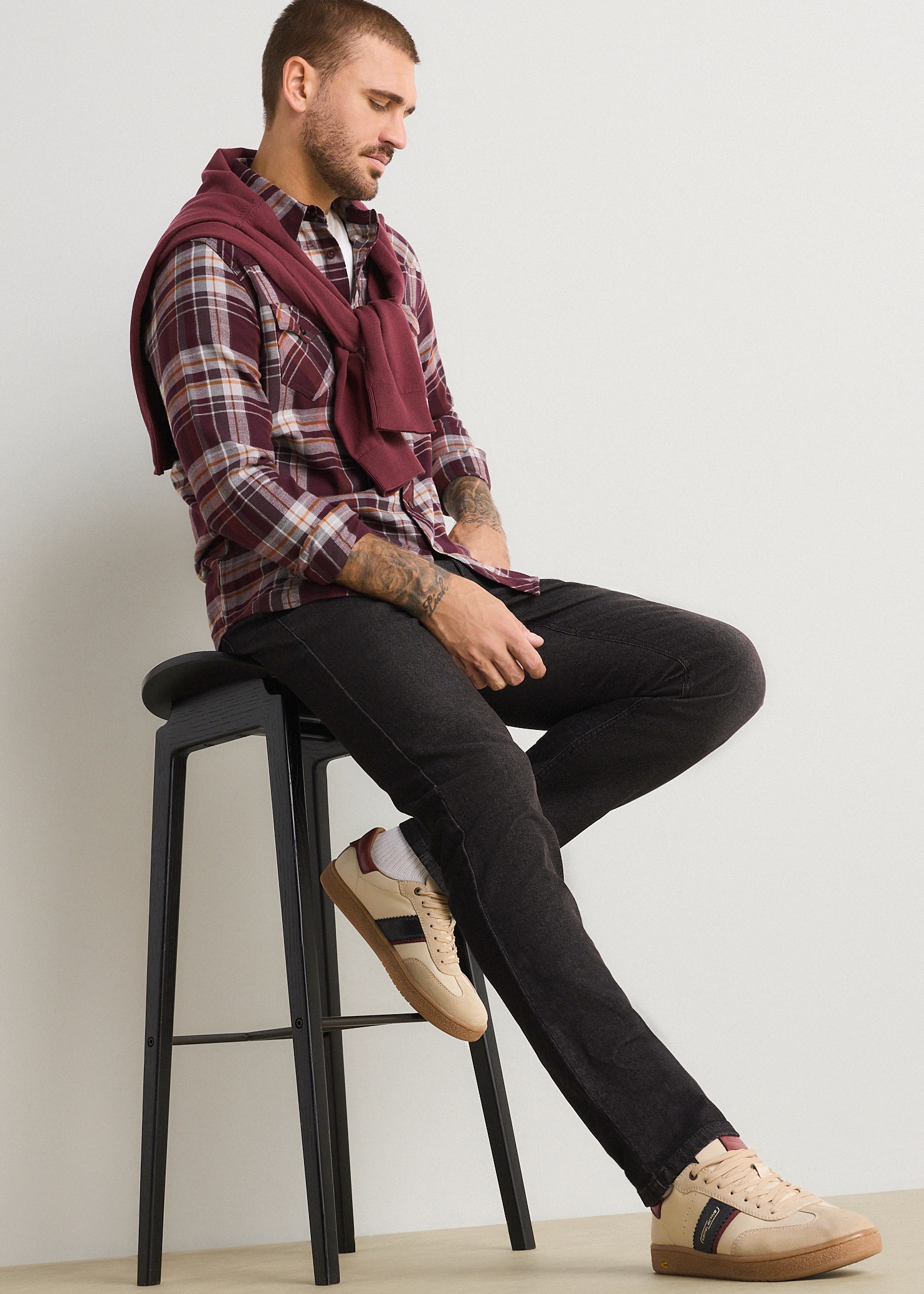 Der Mann trägt ein kariertes Hemd, dunkle Jeans, beige Turnschuhe und einen weinroten Pullover.