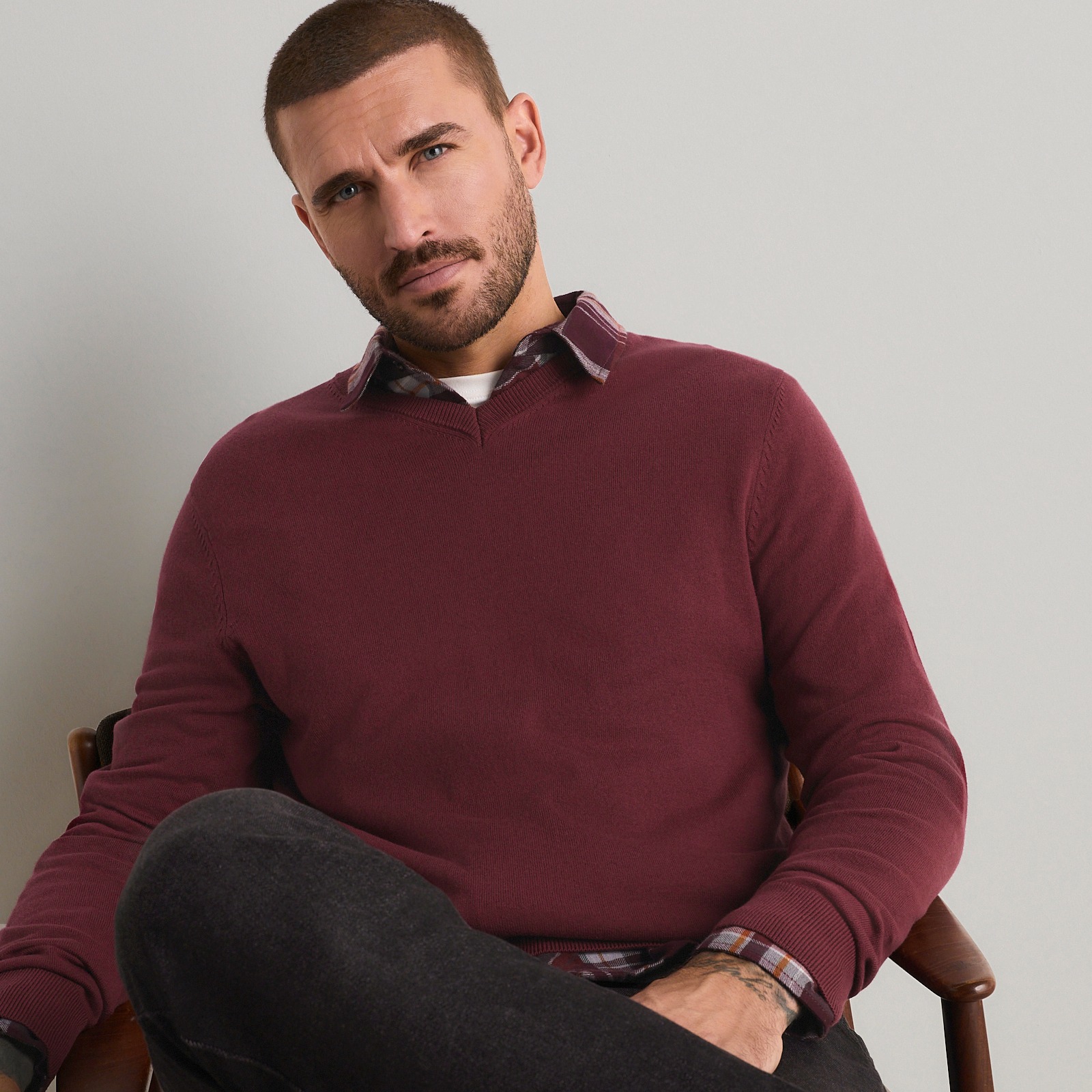 Der Mann trägt einen bordeauxroten Pullover, ein kariertes Hemd und dunkle Jeans.