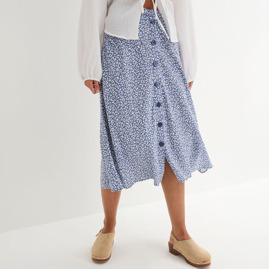 Der gemusterte blaue Rock mit Knöpfen passt perfekt zur weißen Bluse und den beigen Clogs.
