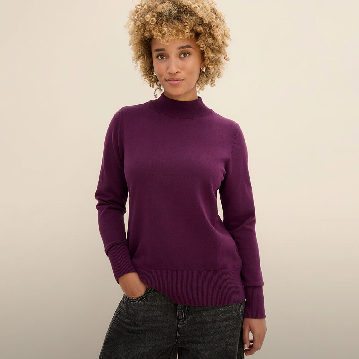 Die Frau trägt einen modischen, lila Pullover mit Stehkragen und dunkle Jeans.