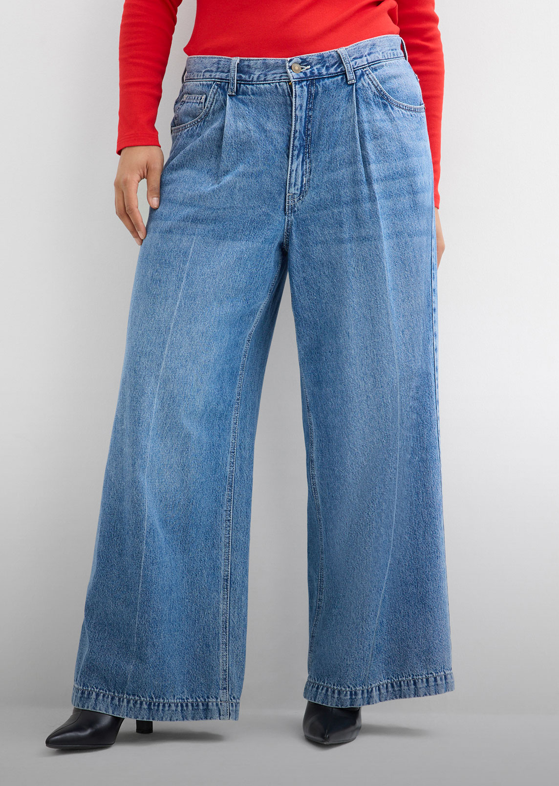 Die Person trägt ein rotes Oberteil, eine weite blaue Bundfaltenjeans und schwarze Pumps.