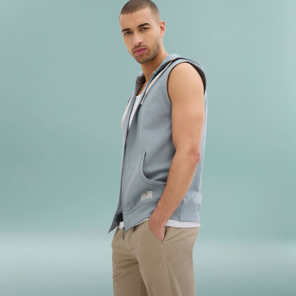 Mann trägt hellblaue Sweatweste mit Kapuze über weißem Shirt und beige Hose.