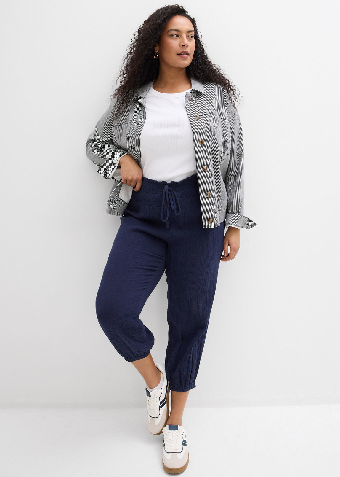 Graue Jacke über weißem Top, dunkelblaue Hose und weiße Sneakers.