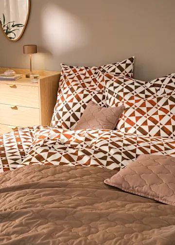 Gemusterte Bettwäsche in Braun- und Orangetönen, dazu Steppdecke und Zierkissen.