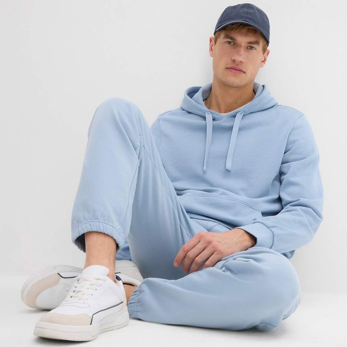 Kapuzenpullover und Jogginghose. Dazu weiße Sneaker und dunkle Baseballkappe.