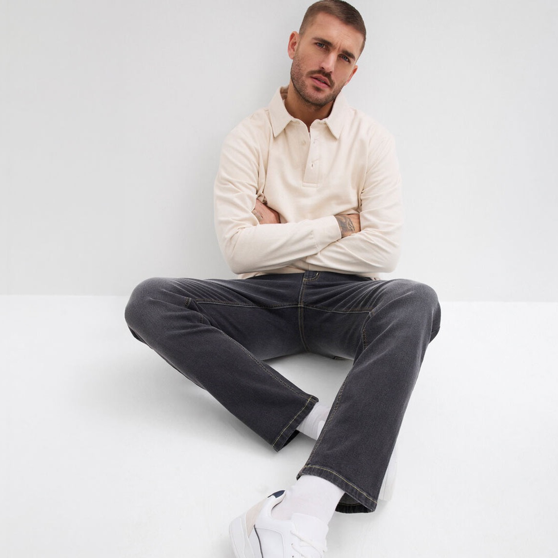 Mann trägt cremefarbenes Shirt, dunkelgraue Hose und weiße Sneaker.