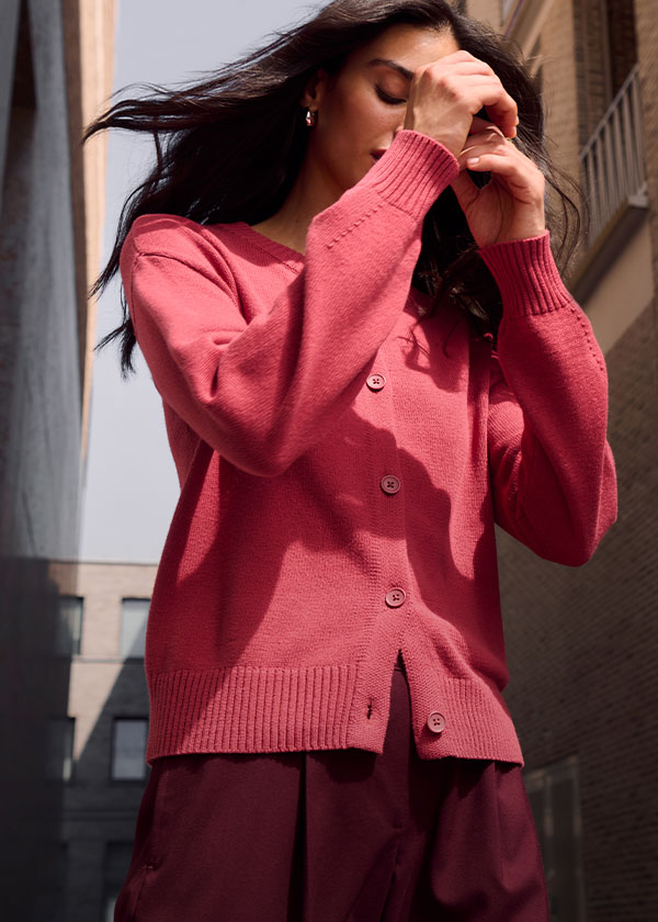 Sie trägt eine leuchtend korallefarbene Strickjacke und eine weinrote Hose.