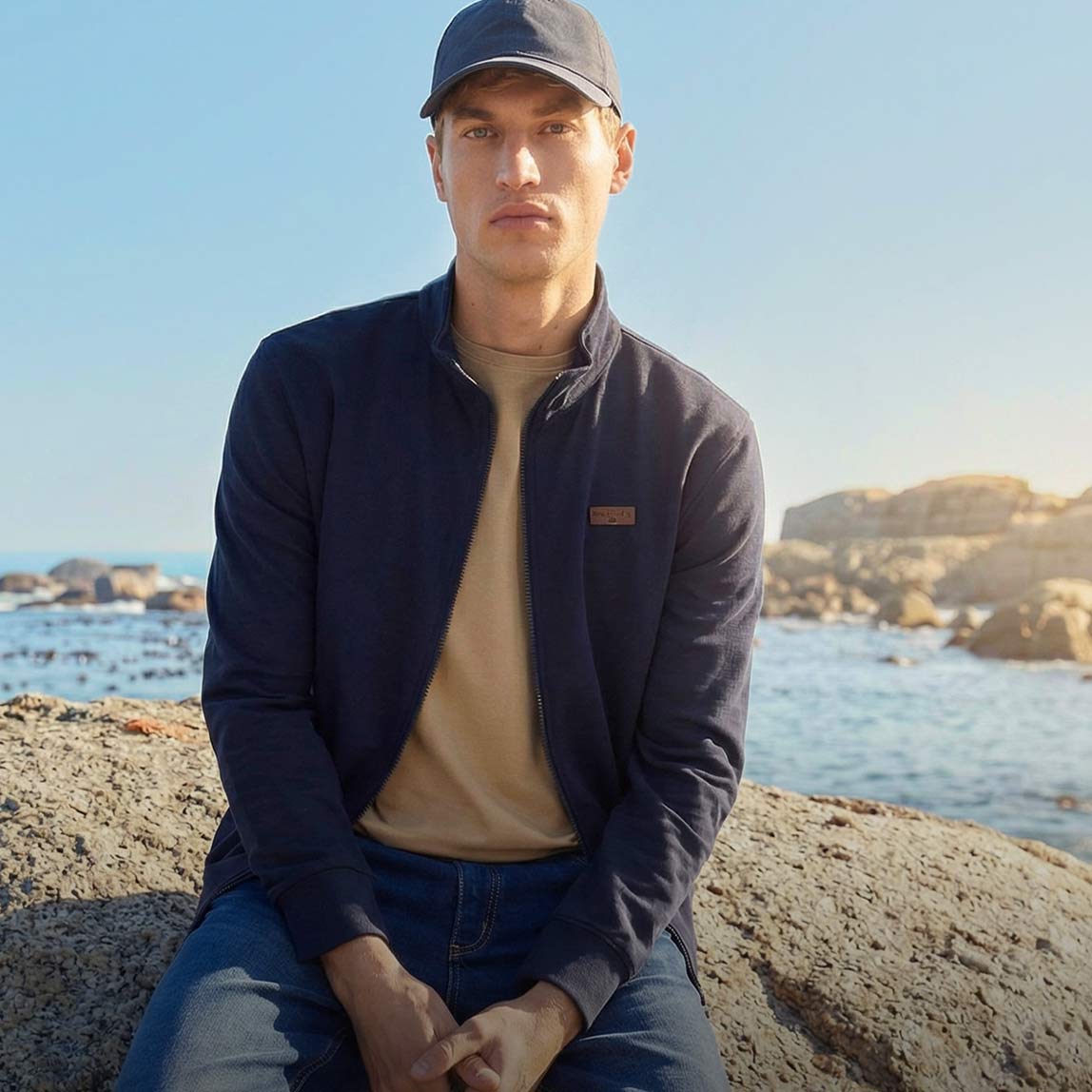 Dunkelblaue Jacke, beiges Shirt, Jeans und Baseballcap am Meer.