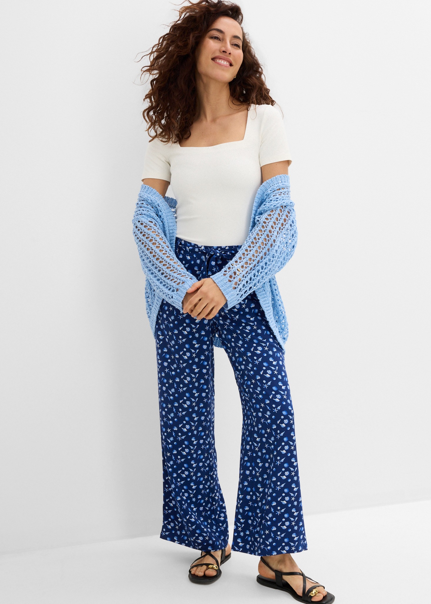 Weißes Oberteil, blaue Musterhose, hellblaue Strickjacke und schwarze Sandalen.