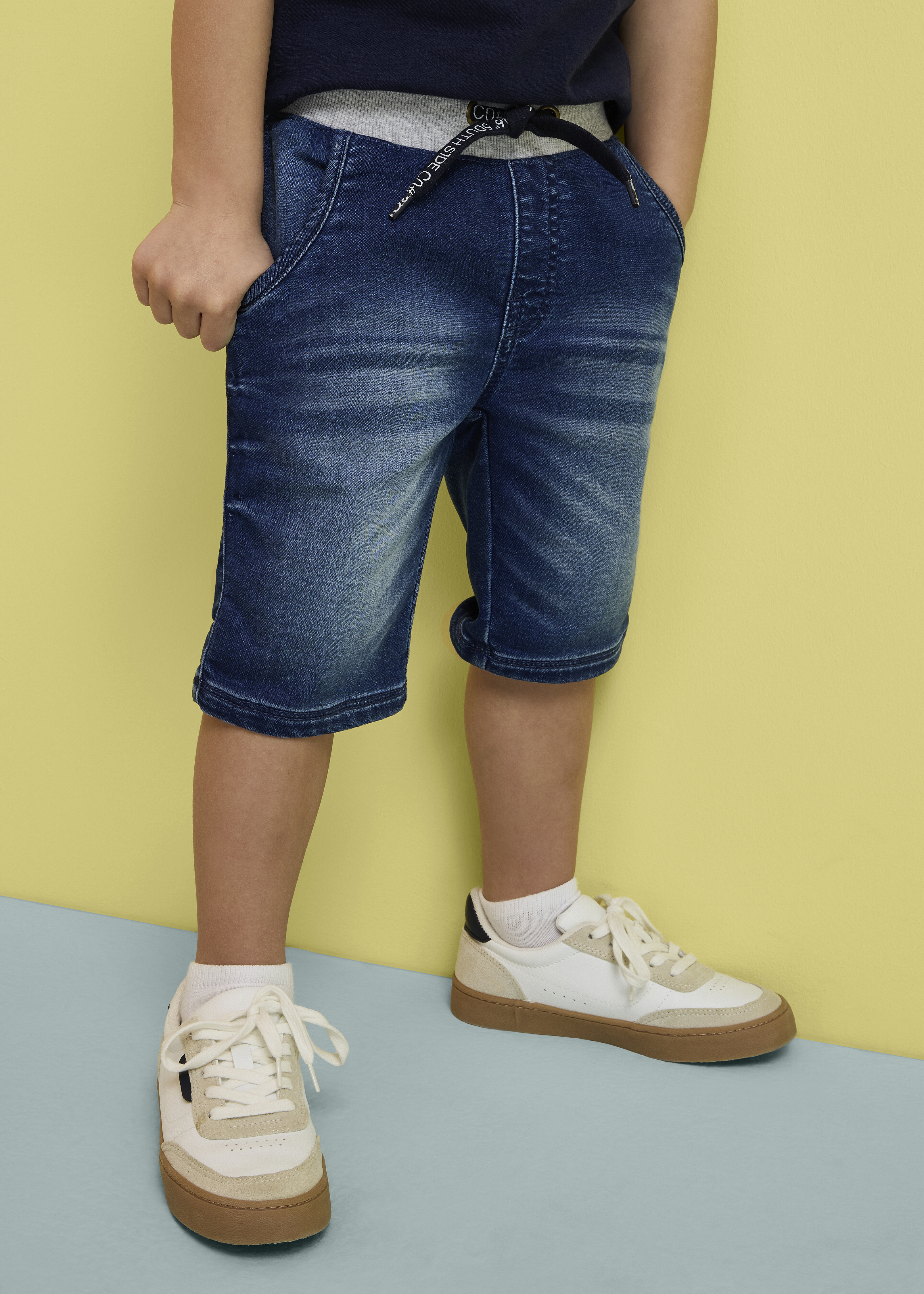 Kind trägt blaue Jeansshorts und weiße Sneaker.