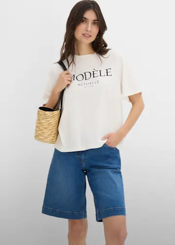 Sie trägt ein lässiges weißes T-Shirt mit Schriftzug und weite blaue Jeans-Bermudas.