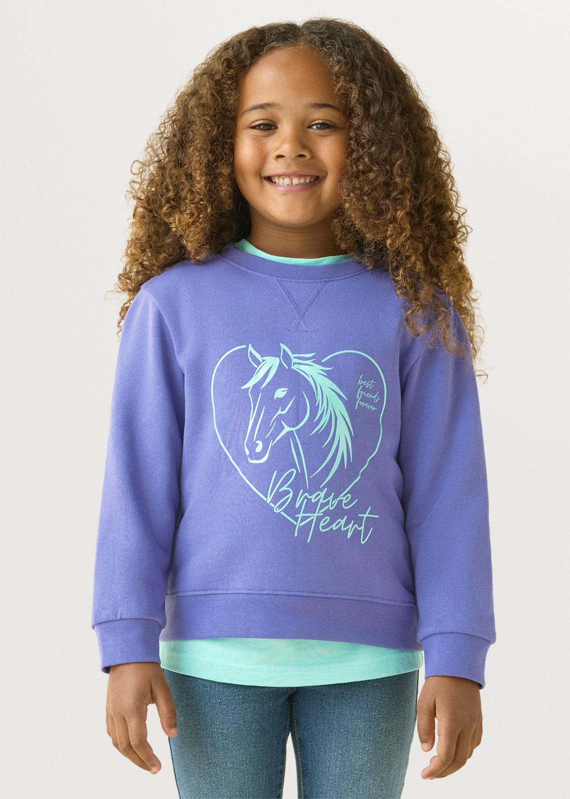 Sie trägt ein lila Sweatshirt mit Pferde-Herz-Motiv, ein hellblaues Shirt und Jeans.