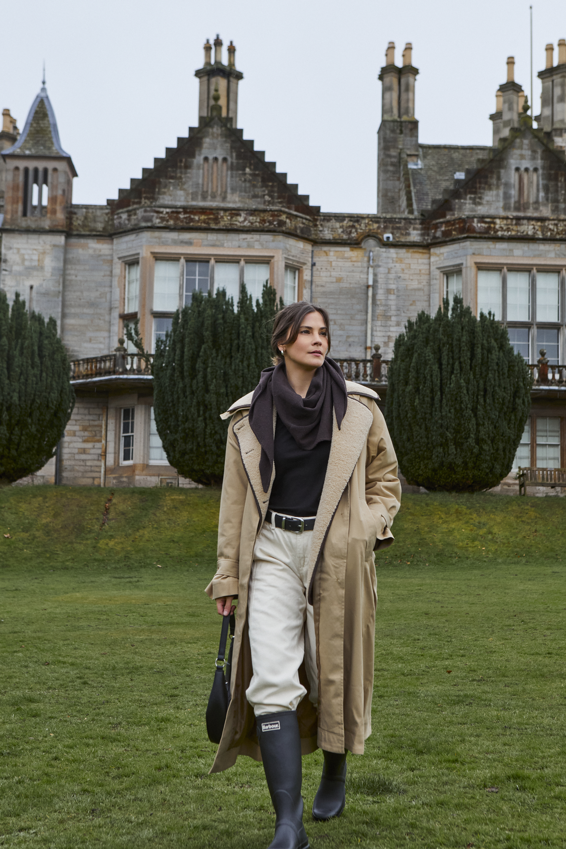 Eleganter beiger Mantel, schwarzer Schal, weiße Hose und Gummistiefel.