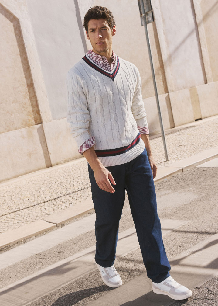 Weißer Zopfmusterpullover über Hemd, dunkle Hose und weiße Sneakers.