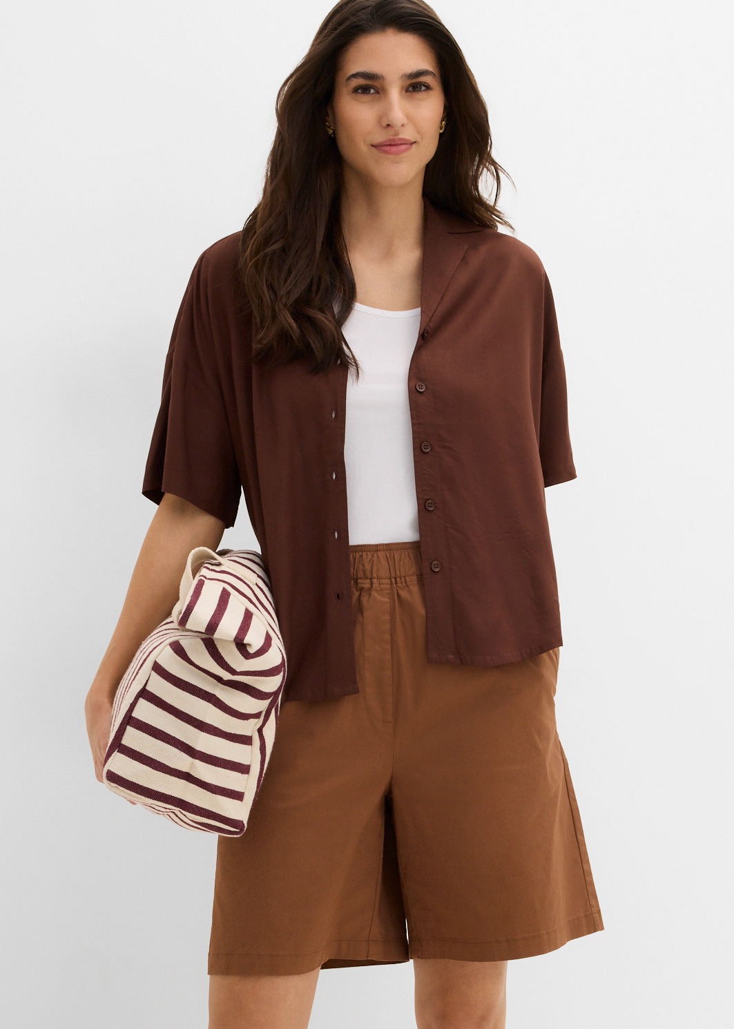 Braunes Overshirt über weißem Top, kombiniert mit bequemen Shorts und gestreifter Tasche.